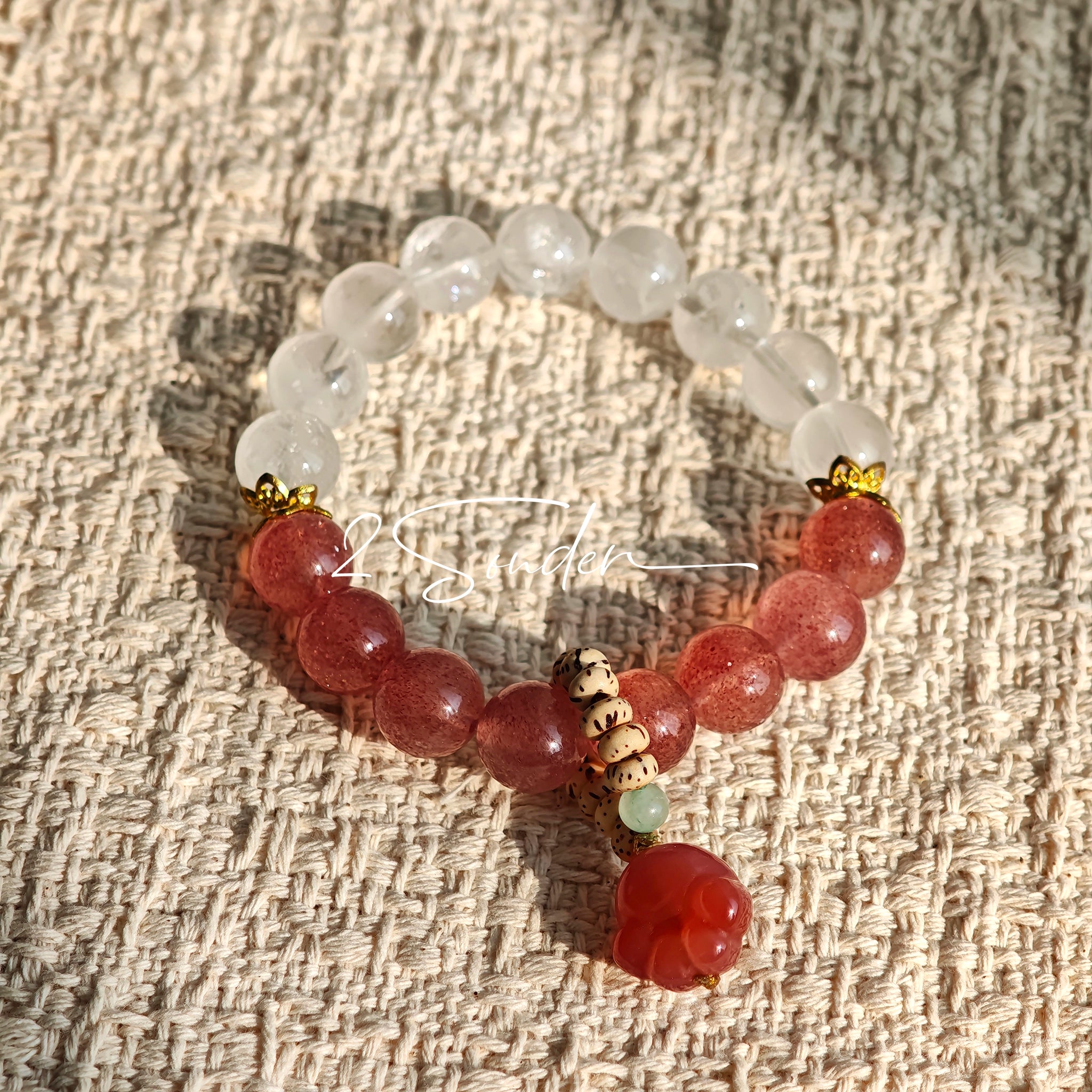 This bracelet features a beautiful blend of translucent white phantom quartz beads and rich pink strawberry quartz beads, creating a striking contrast. Adorned with delicate gold-toned accents and a charming red cat's paw charm, it exudes a sweet and elegant vibe. The combination of smooth crystal spheres and a playful pendant makes it a lovely and eye-catching accessory.