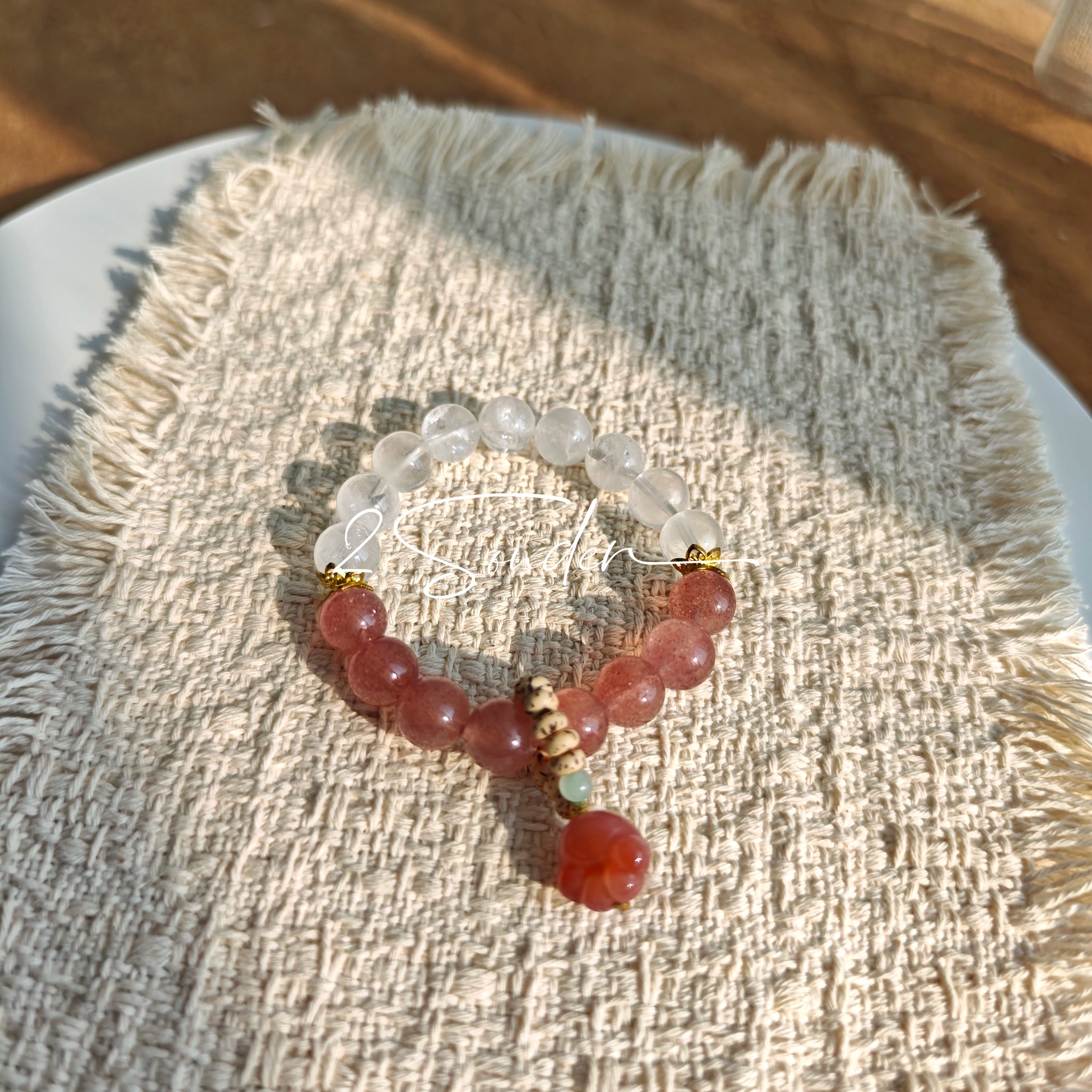 This bracelet features a beautiful blend of translucent white phantom quartz beads and rich pink strawberry quartz beads, creating a striking contrast. Adorned with delicate gold-toned accents and a charming red cat's paw charm, it exudes a sweet and elegant vibe. The combination of smooth crystal spheres and a playful pendant makes it a lovely and eye-catching accessory.