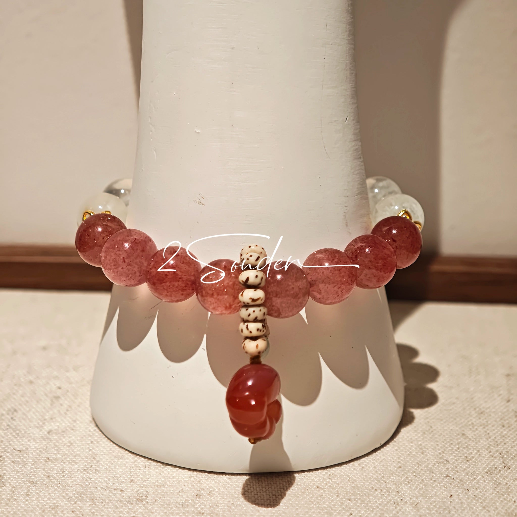 This bracelet features a beautiful blend of translucent white phantom quartz beads and rich pink strawberry quartz beads, creating a striking contrast. Adorned with delicate gold-toned accents and a charming red cat's paw charm, it exudes a sweet and elegant vibe. The combination of smooth crystal spheres and a playful pendant makes it a lovely and eye-catching accessory.