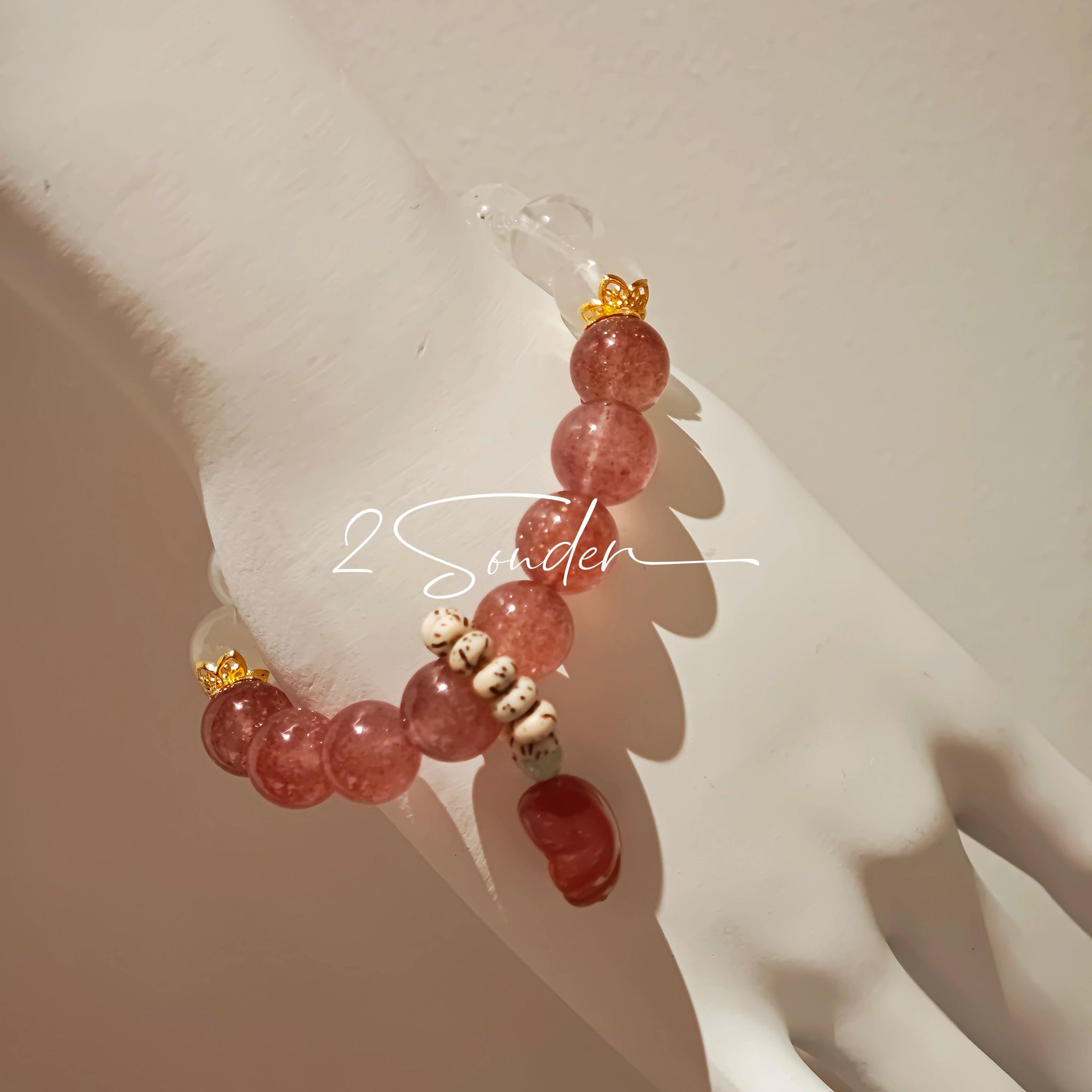 This bracelet features a beautiful blend of translucent white phantom quartz beads and rich pink strawberry quartz beads, creating a striking contrast. Adorned with delicate gold-toned accents and a charming red cat's paw charm, it exudes a sweet and elegant vibe. The combination of smooth crystal spheres and a playful pendant makes it a lovely and eye-catching accessory.