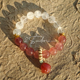 This bracelet features a beautiful blend of translucent white phantom quartz beads and rich pink strawberry quartz beads, creating a striking contrast. Adorned with delicate gold-toned accents and a charming red cat's paw charm, it exudes a sweet and elegant vibe. The combination of smooth crystal spheres and a playful pendant makes it a lovely and eye-catching accessory.