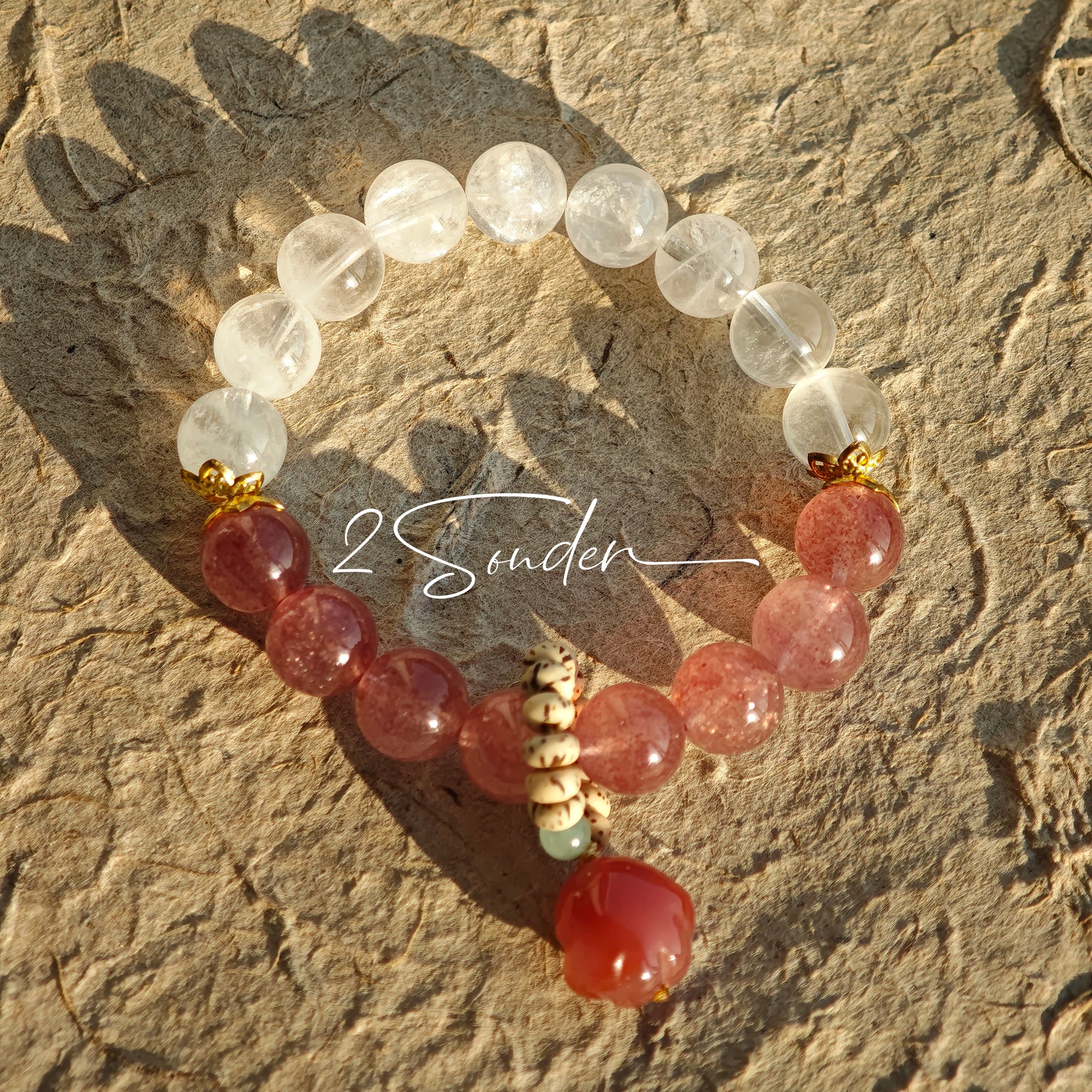 This bracelet features a beautiful blend of translucent white phantom quartz beads and rich pink strawberry quartz beads, creating a striking contrast. Adorned with delicate gold-toned accents and a charming red cat's paw charm, it exudes a sweet and elegant vibe. The combination of smooth crystal spheres and a playful pendant makes it a lovely and eye-catching accessory.