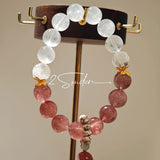 This bracelet features a beautiful blend of translucent white phantom quartz beads and rich pink strawberry quartz beads, creating a striking contrast. Adorned with delicate gold-toned accents and a charming red cat's paw charm, it exudes a sweet and elegant vibe. The combination of smooth crystal spheres and a playful pendant makes it a lovely and eye-catching accessory.