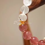 This bracelet features a beautiful blend of translucent white phantom quartz beads and rich pink strawberry quartz beads, creating a striking contrast. Adorned with delicate gold-toned accents and a charming red cat's paw charm, it exudes a sweet and elegant vibe. The combination of smooth crystal spheres and a playful pendant makes it a lovely and eye-catching accessory.