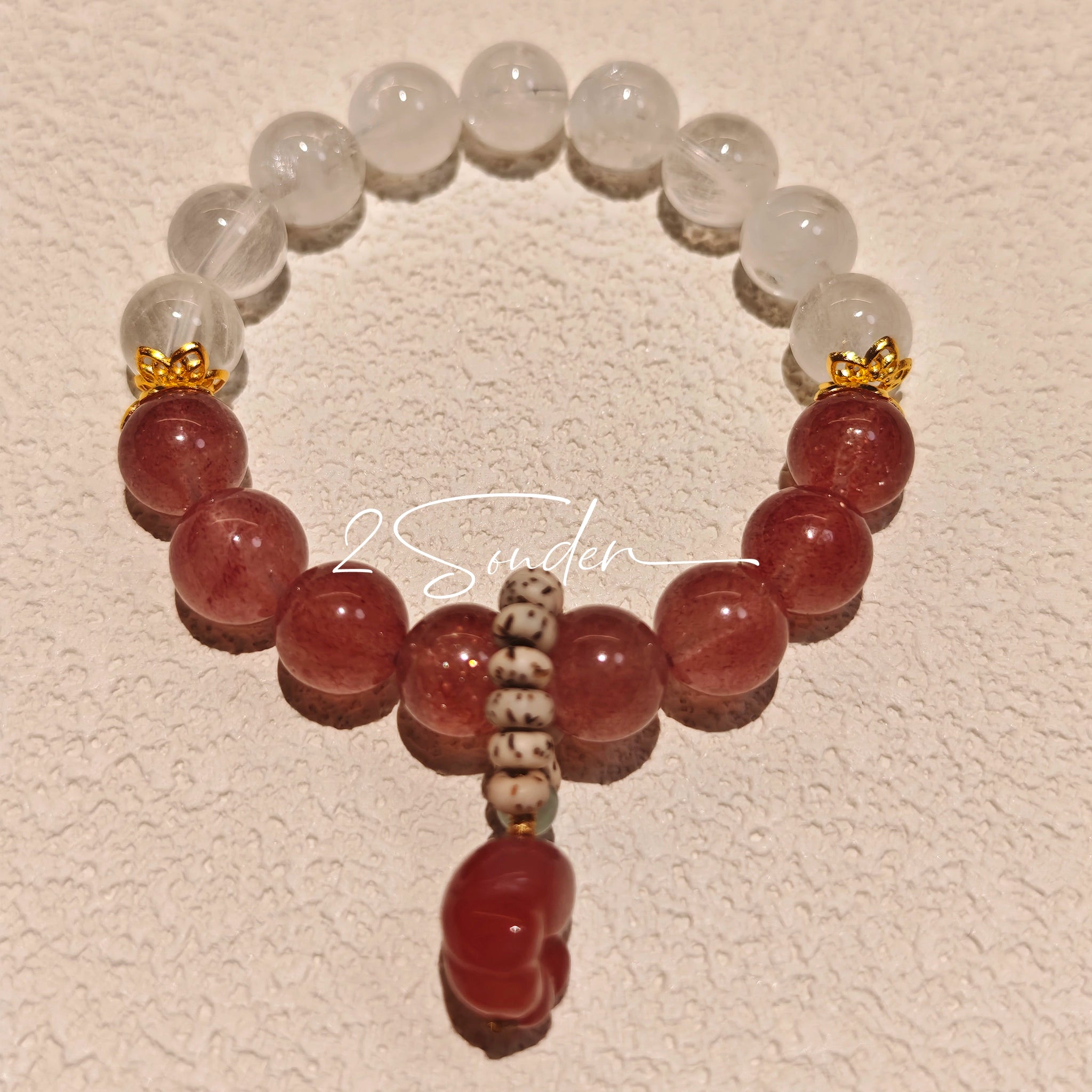 This bracelet features a beautiful blend of translucent white phantom quartz beads and rich pink strawberry quartz beads, creating a striking contrast. Adorned with delicate gold-toned accents and a charming red cat's paw charm, it exudes a sweet and elegant vibe. The combination of smooth crystal spheres and a playful pendant makes it a lovely and eye-catching accessory.