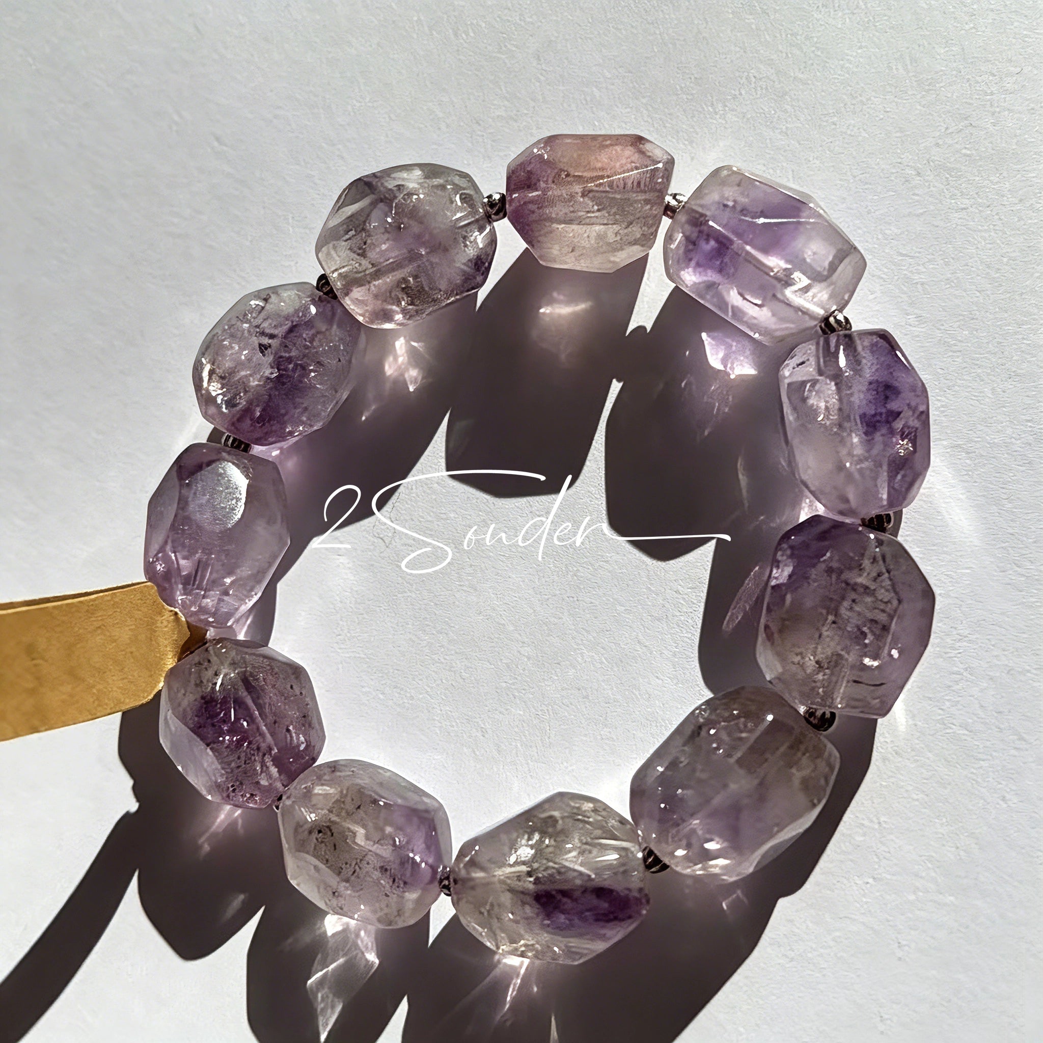 This exquisite Super Seven bracelet showcases irregularly faceted beads in a captivating blend of soft lavender and smoky purple hues, each displaying unique natural inclusions that catch the light beautifully. The polished crystals are seamlessly interspersed with tiny metallic spacer beads, culminating in an ornate silver-tone decorative bead that adds a touch of elegance to the organic design. Crafted with meticulous attention to detail, the bracelet offers a harmonious blend of rare gemstone texture and