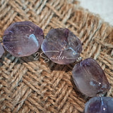 This exquisite Super Seven bracelet showcases irregularly faceted beads in a captivating blend of soft lavender and smoky purple hues, each displaying unique natural inclusions that catch the light beautifully. The polished crystals are seamlessly interspersed with tiny metallic spacer beads, culminating in an ornate silver-tone decorative bead that adds a touch of elegance to the organic design. Crafted with meticulous attention to detail, the bracelet offers a harmonious blend of rare gemstone texture and