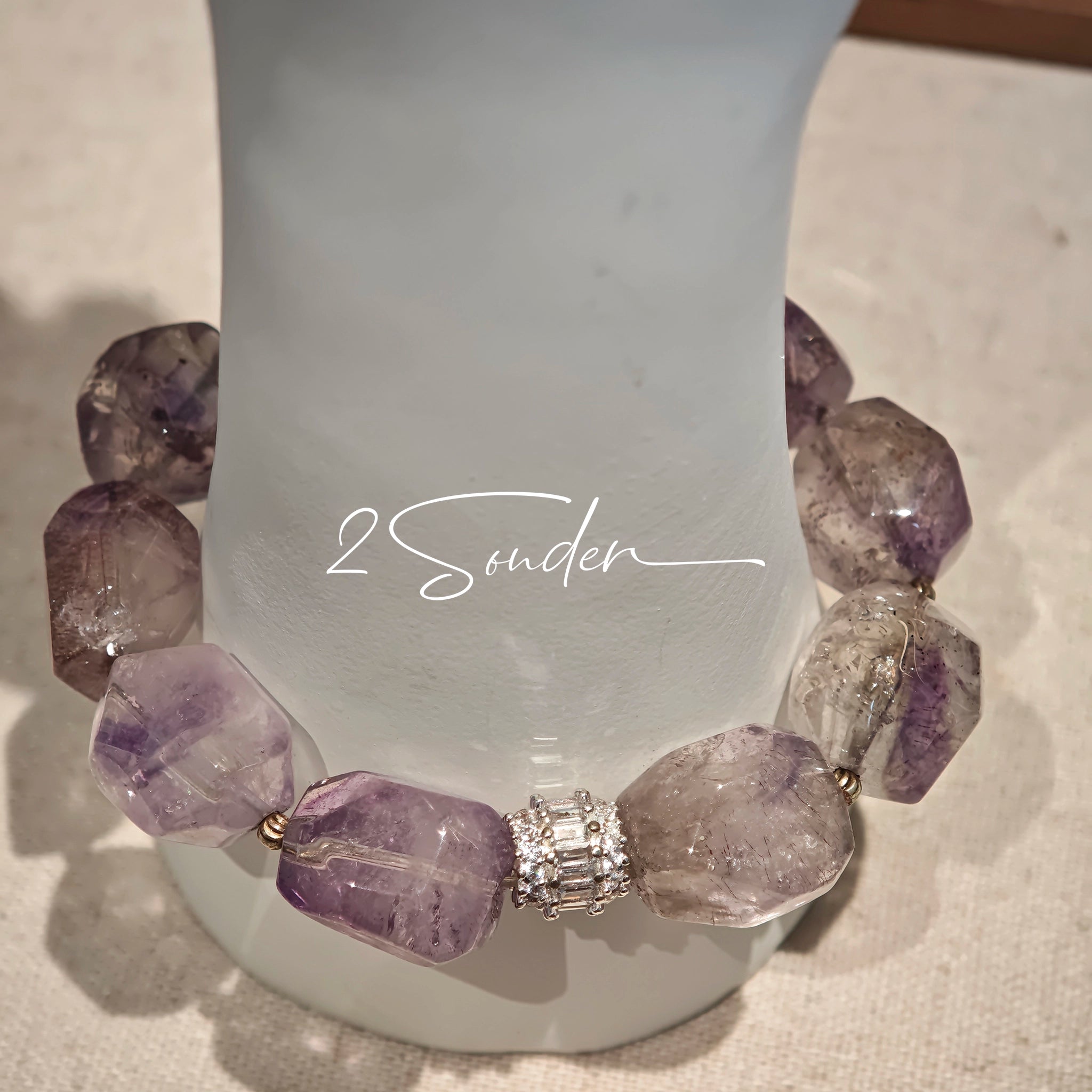 This exquisite Super Seven bracelet showcases irregularly faceted beads in a captivating blend of soft lavender and smoky purple hues, each displaying unique natural inclusions that catch the light beautifully. The polished crystals are seamlessly interspersed with tiny metallic spacer beads, culminating in an ornate silver-tone decorative bead that adds a touch of elegance to the organic design. Crafted with meticulous attention to detail, the bracelet offers a harmonious blend of rare gemstone texture and