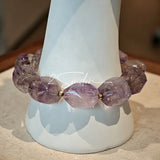 This exquisite Super Seven bracelet showcases irregularly faceted beads in a captivating blend of soft lavender and smoky purple hues, each displaying unique natural inclusions that catch the light beautifully. The polished crystals are seamlessly interspersed with tiny metallic spacer beads, culminating in an ornate silver-tone decorative bead that adds a touch of elegance to the organic design. Crafted with meticulous attention to detail, the bracelet offers a harmonious blend of rare gemstone texture and