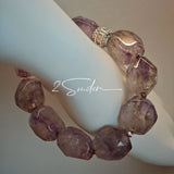 This exquisite Super Seven bracelet showcases irregularly faceted beads in a captivating blend of soft lavender and smoky purple hues, each displaying unique natural inclusions that catch the light beautifully. The polished crystals are seamlessly interspersed with tiny metallic spacer beads, culminating in an ornate silver-tone decorative bead that adds a touch of elegance to the organic design. Crafted with meticulous attention to detail, the bracelet offers a harmonious blend of rare gemstone texture and