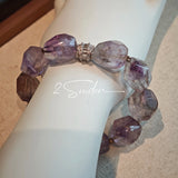 This exquisite Super Seven bracelet showcases irregularly faceted beads in a captivating blend of soft lavender and smoky purple hues, each displaying unique natural inclusions that catch the light beautifully. The polished crystals are seamlessly interspersed with tiny metallic spacer beads, culminating in an ornate silver-tone decorative bead that adds a touch of elegance to the organic design. Crafted with meticulous attention to detail, the bracelet offers a harmonious blend of rare gemstone texture and