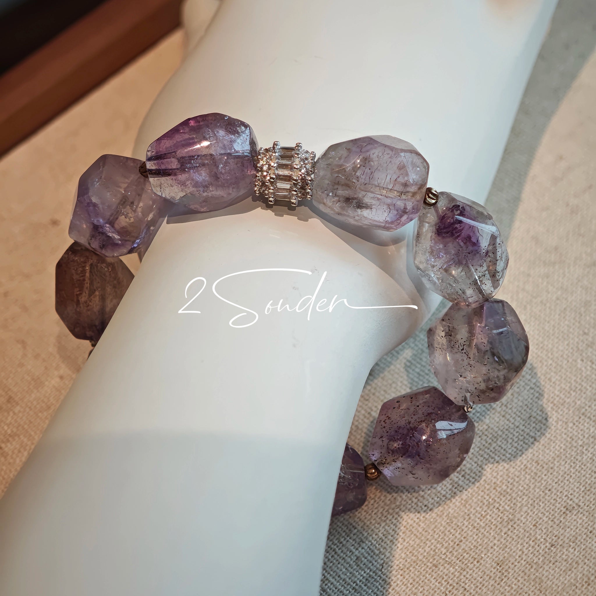 This exquisite Super Seven bracelet showcases irregularly faceted beads in a captivating blend of soft lavender and smoky purple hues, each displaying unique natural inclusions that catch the light beautifully. The polished crystals are seamlessly interspersed with tiny metallic spacer beads, culminating in an ornate silver-tone decorative bead that adds a touch of elegance to the organic design. Crafted with meticulous attention to detail, the bracelet offers a harmonious blend of rare gemstone texture and
