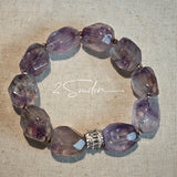 This exquisite Super Seven bracelet showcases irregularly faceted beads in a captivating blend of soft lavender and smoky purple hues, each displaying unique natural inclusions that catch the light beautifully. The polished crystals are seamlessly interspersed with tiny metallic spacer beads, culminating in an ornate silver-tone decorative bead that adds a touch of elegance to the organic design. Crafted with meticulous attention to detail, the bracelet offers a harmonious blend of rare gemstone texture and