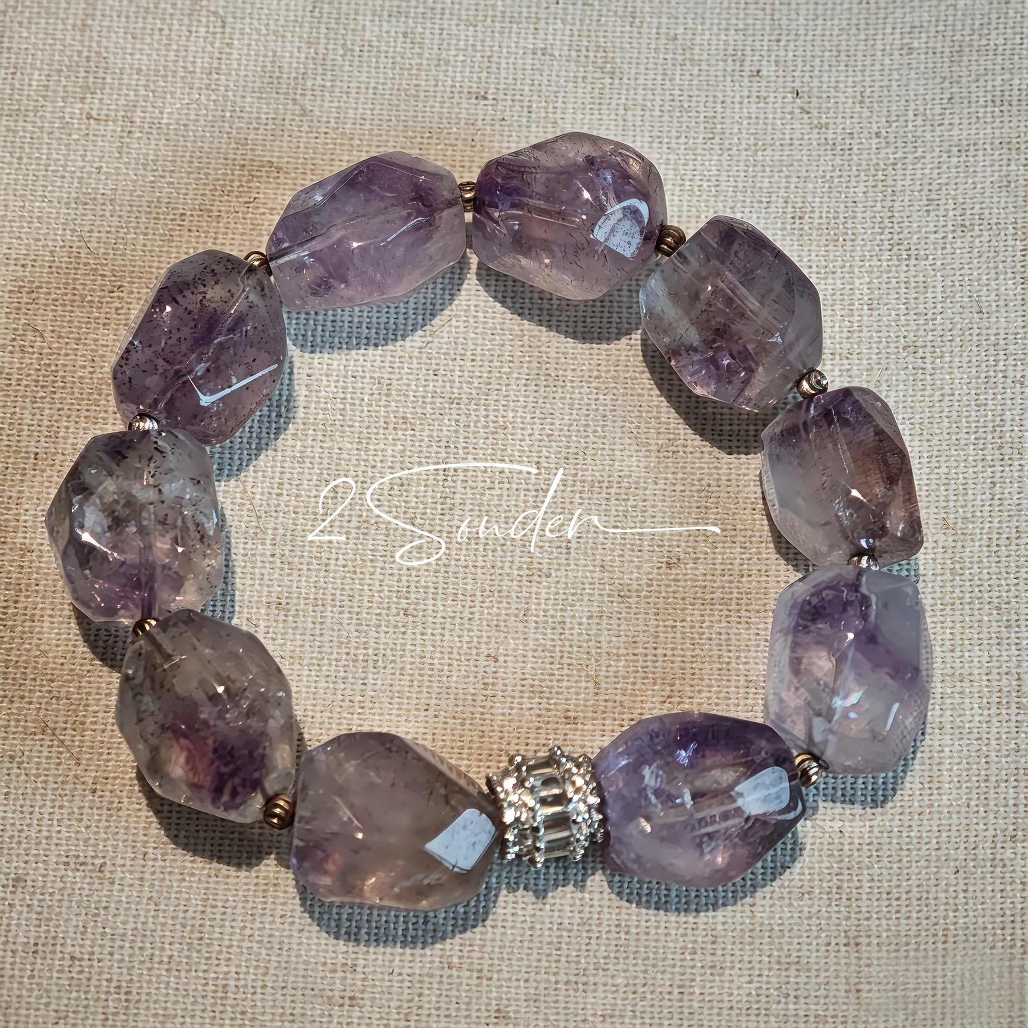 This exquisite Super Seven bracelet showcases irregularly faceted beads in a captivating blend of soft lavender and smoky purple hues, each displaying unique natural inclusions that catch the light beautifully. The polished crystals are seamlessly interspersed with tiny metallic spacer beads, culminating in an ornate silver-tone decorative bead that adds a touch of elegance to the organic design. Crafted with meticulous attention to detail, the bracelet offers a harmonious blend of rare gemstone texture and