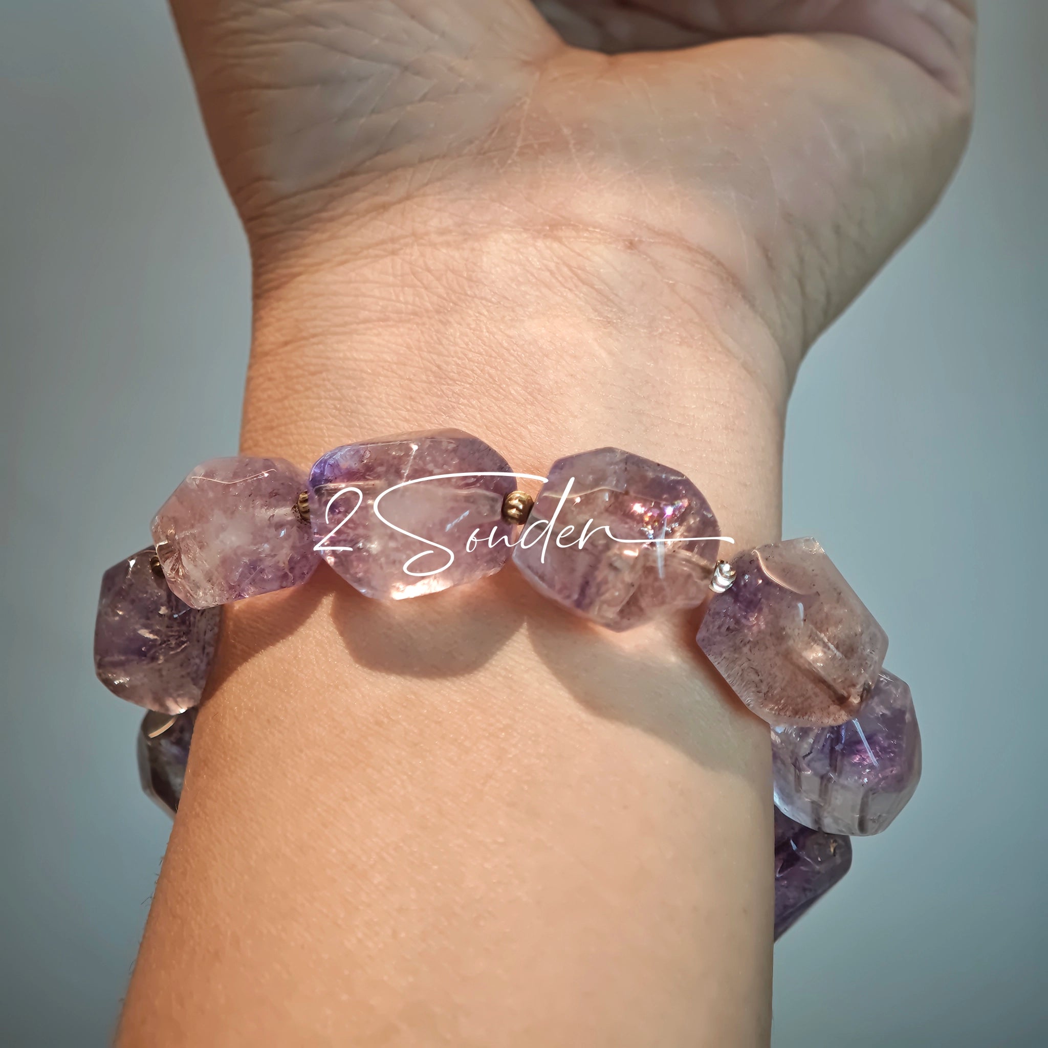 This exquisite Super Seven bracelet showcases irregularly faceted beads in a captivating blend of soft lavender and smoky purple hues, each displaying unique natural inclusions that catch the light beautifully. The polished crystals are seamlessly interspersed with tiny metallic spacer beads, culminating in an ornate silver-tone decorative bead that adds a touch of elegance to the organic design. Crafted with meticulous attention to detail, the bracelet offers a harmonious blend of rare gemstone texture and