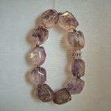 This exquisite Super Seven bracelet showcases irregularly faceted beads in a captivating blend of soft lavender and smoky purple hues, each displaying unique natural inclusions that catch the light beautifully. The polished crystals are seamlessly interspersed with tiny metallic spacer beads, culminating in an ornate silver-tone decorative bead that adds a touch of elegance to the organic design. Crafted with meticulous attention to detail, the bracelet offers a harmonious blend of rare gemstone texture and