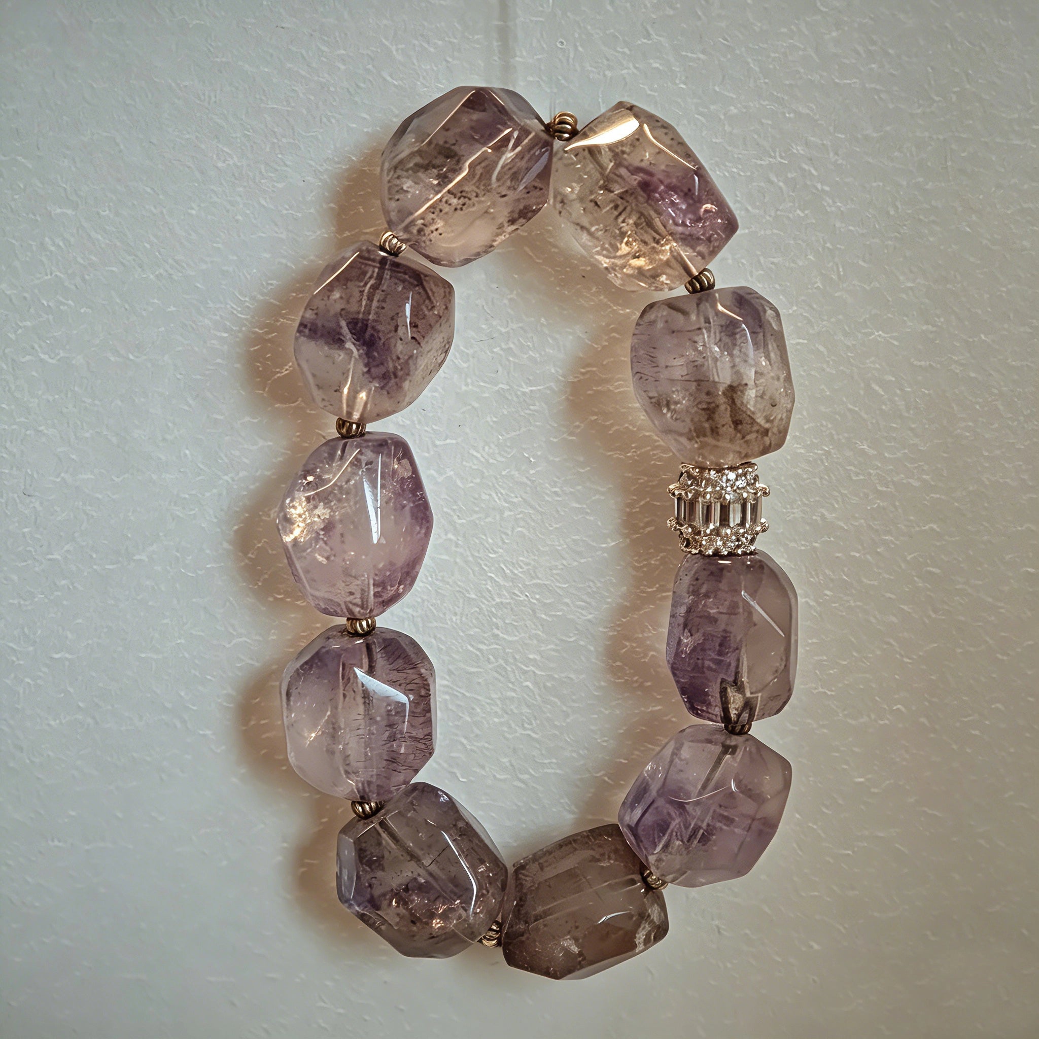 This exquisite Super Seven bracelet showcases irregularly faceted beads in a captivating blend of soft lavender and smoky purple hues, each displaying unique natural inclusions that catch the light beautifully. The polished crystals are seamlessly interspersed with tiny metallic spacer beads, culminating in an ornate silver-tone decorative bead that adds a touch of elegance to the organic design. Crafted with meticulous attention to detail, the bracelet offers a harmonious blend of rare gemstone texture and