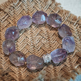 This exquisite Super Seven bracelet showcases irregularly faceted beads in a captivating blend of soft lavender and smoky purple hues, each displaying unique natural inclusions that catch the light beautifully. The polished crystals are seamlessly interspersed with tiny metallic spacer beads, culminating in an ornate silver-tone decorative bead that adds a touch of elegance to the organic design. Crafted with meticulous attention to detail, the bracelet offers a harmonious blend of rare gemstone texture and