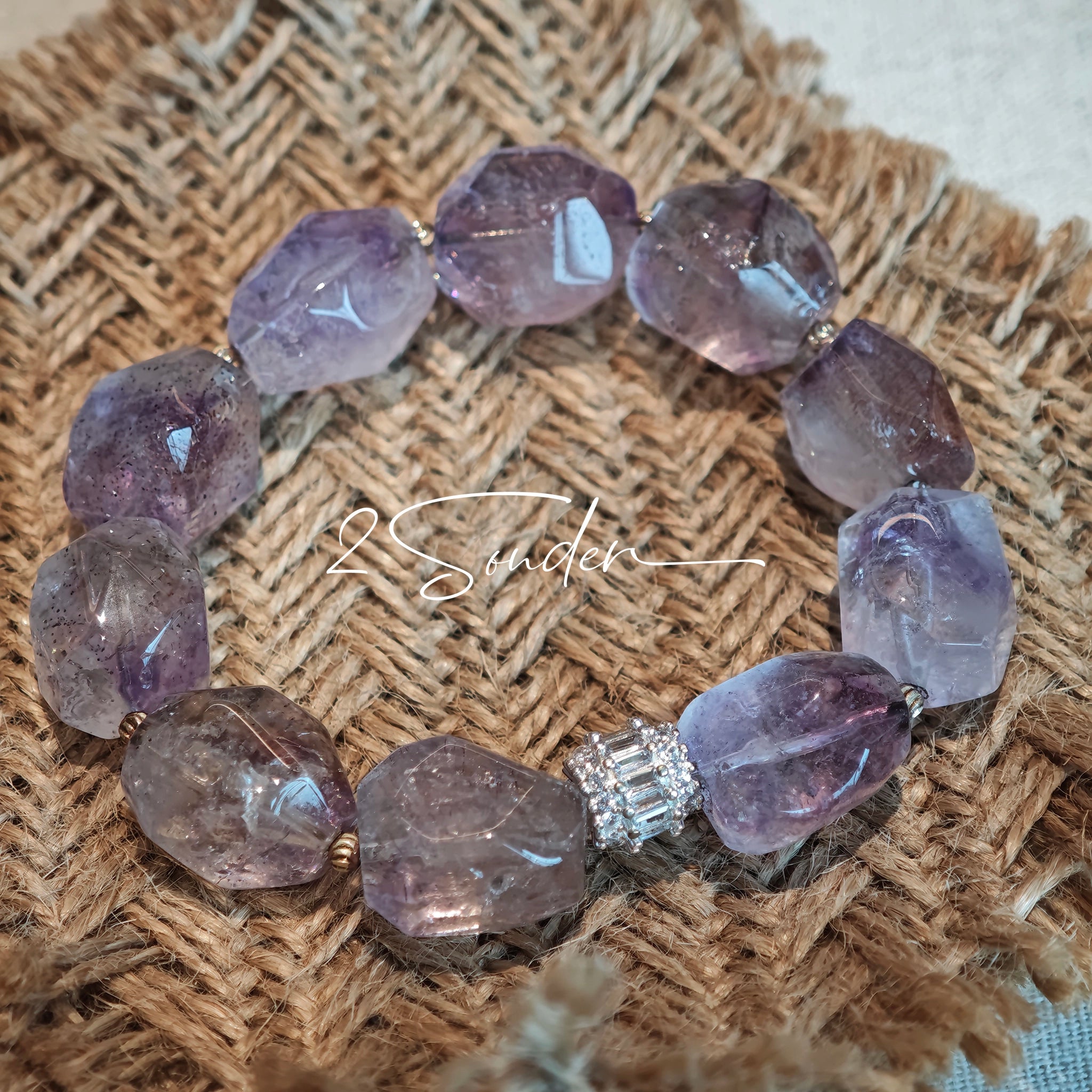 This exquisite Super Seven bracelet showcases irregularly faceted beads in a captivating blend of soft lavender and smoky purple hues, each displaying unique natural inclusions that catch the light beautifully. The polished crystals are seamlessly interspersed with tiny metallic spacer beads, culminating in an ornate silver-tone decorative bead that adds a touch of elegance to the organic design. Crafted with meticulous attention to detail, the bracelet offers a harmonious blend of rare gemstone texture and