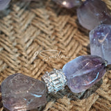 This exquisite Super Seven bracelet showcases irregularly faceted beads in a captivating blend of soft lavender and smoky purple hues, each displaying unique natural inclusions that catch the light beautifully. The polished crystals are seamlessly interspersed with tiny metallic spacer beads, culminating in an ornate silver-tone decorative bead that adds a touch of elegance to the organic design. Crafted with meticulous attention to detail, the bracelet offers a harmonious blend of rare gemstone texture and
