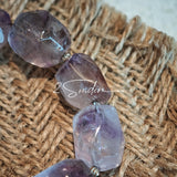 This exquisite Super Seven bracelet showcases irregularly faceted beads in a captivating blend of soft lavender and smoky purple hues, each displaying unique natural inclusions that catch the light beautifully. The polished crystals are seamlessly interspersed with tiny metallic spacer beads, culminating in an ornate silver-tone decorative bead that adds a touch of elegance to the organic design. Crafted with meticulous attention to detail, the bracelet offers a harmonious blend of rare gemstone texture and