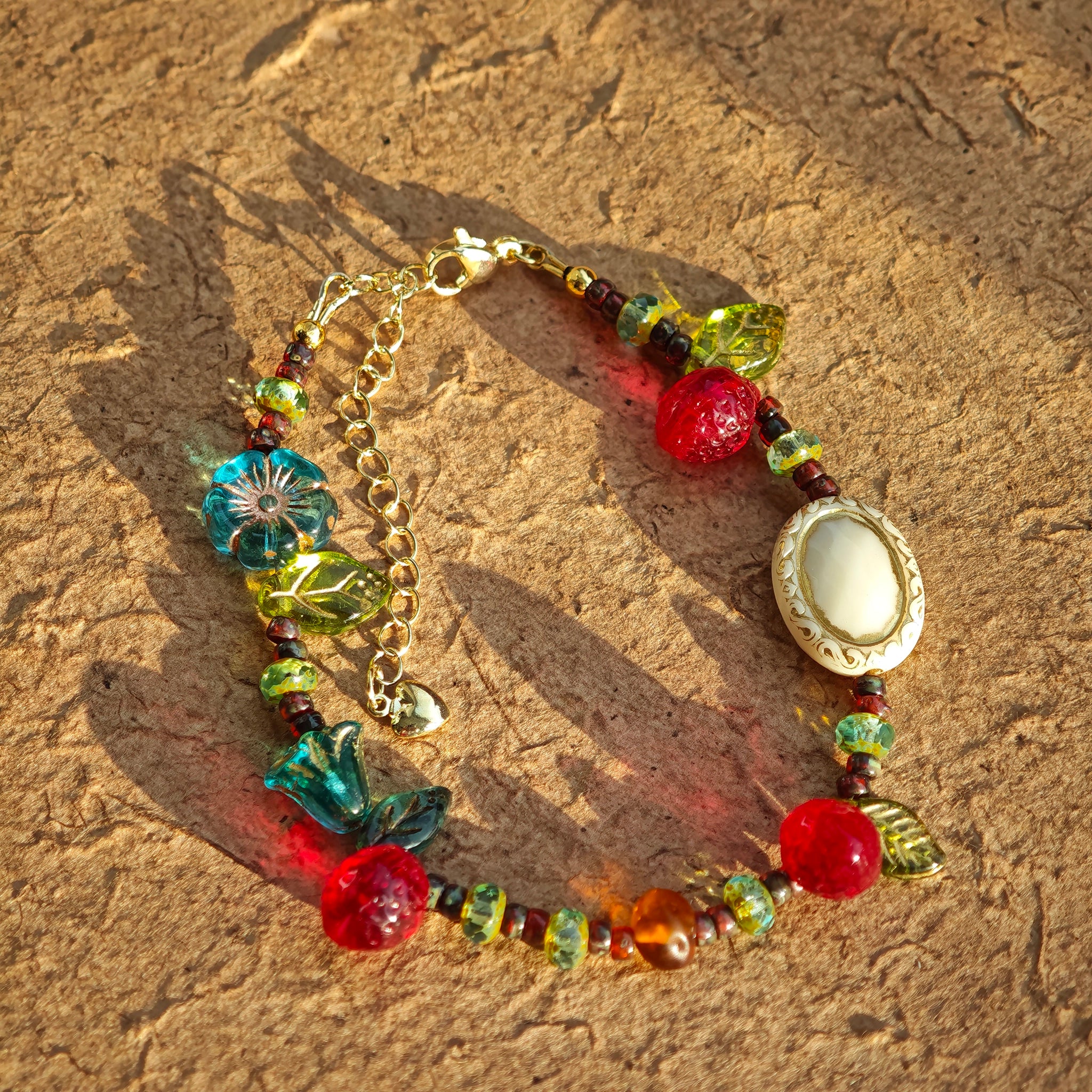 This vibrant bracelet features a colorful mix of ruby-red lemon, sapphire-blue flower, and emerald-green leaf charms, all dangling from a shimmering Czech glass bead chain.