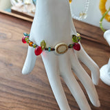 This vibrant bracelet features a colorful mix of ruby-red lemon, sapphire-blue flower, and emerald-green leaf charms, all dangling from a shimmering Czech glass bead chain.