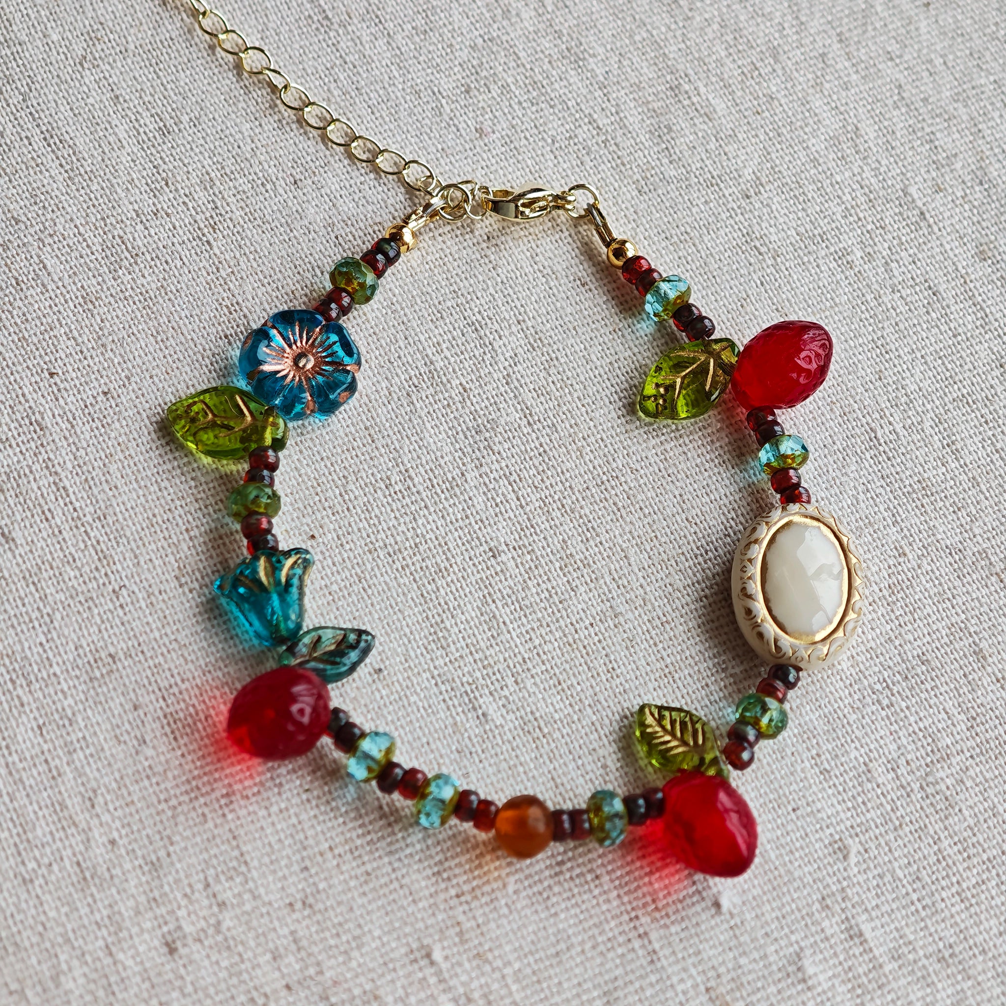 This vibrant bracelet features a colorful mix of ruby-red lemon, sapphire-blue flower, and emerald-green leaf charms, all dangling from a shimmering Czech glass bead chain.