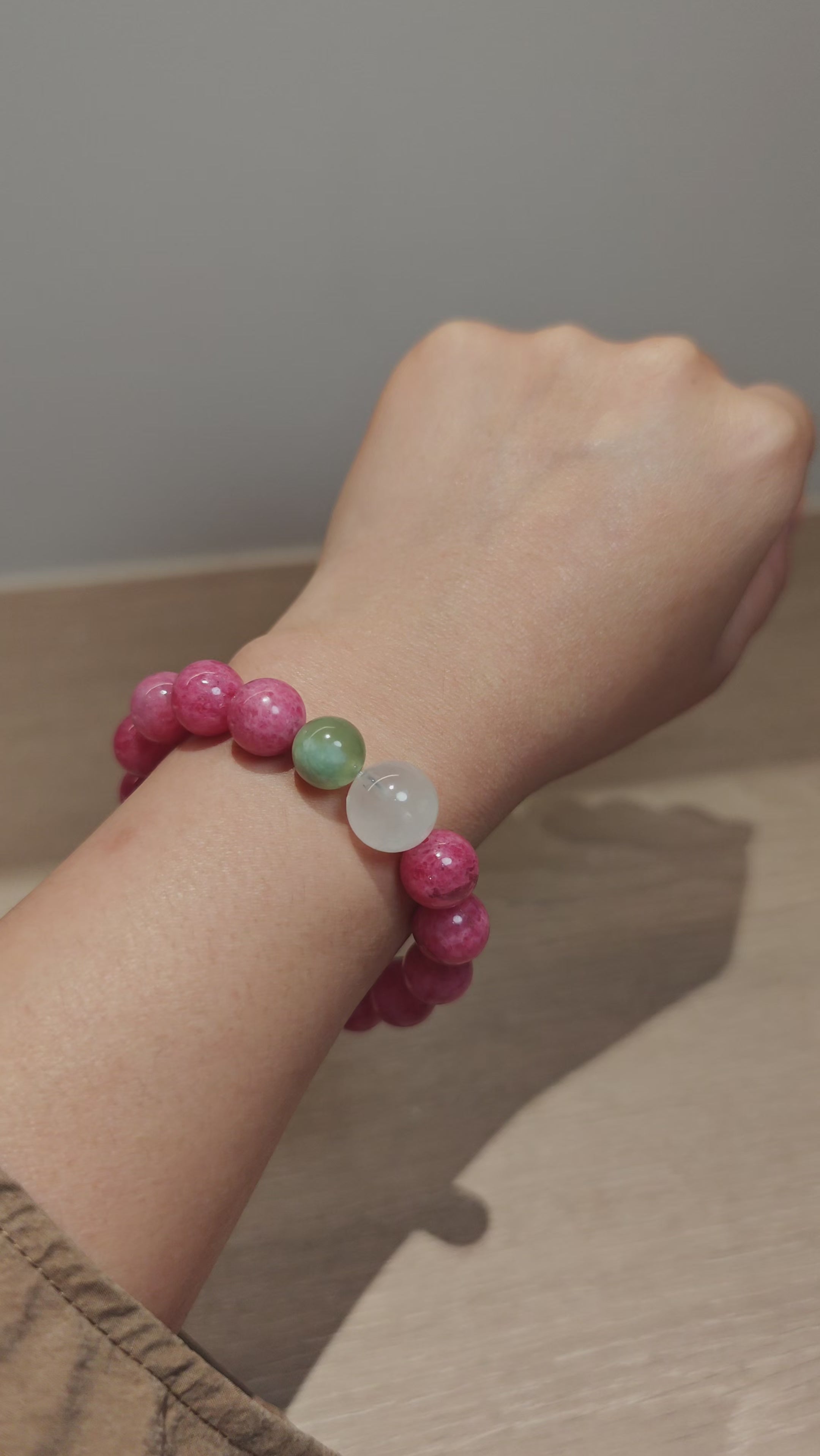 This beaded bracelet is mainly made of round, rosy-pink rhodonite beads with a delicate marbled texture. It is accented by one pale green Xiuyan jade bead (in the "green grape milk cap" style) and one translucent white phantom quartz bead, creating a soft color contrast against the dominant pink hue. The smooth, polished stones give the bracelet a gentle, natural look.