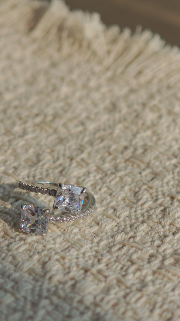 This striking bypass ring showcases two large, brilliant cushion-cut clear stones held securely in classic four-prong settings. The elegant twisted band is lined with a row of sparkling accent Cubic Zirconia stones, delivering a high-end, light luxury aesthetic that instantly catches the eye.