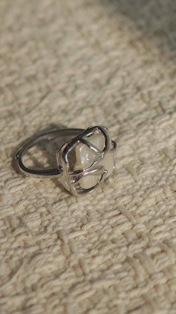 This adjustable S925 sterling silver ring features a striking baroque pearl at its center, set within an organic, flowing silver frame that wraps around the irregular, lustrous gemstone. The open, sculptural metalwork creates a modern, artistic silhouette, showcasing the pearl's natural, one-of-a-kind texture and soft iridescence. The sleek, polished silver band complements the unique pearl, making it a standout piece with a contemporary, bohemian aesthetic.