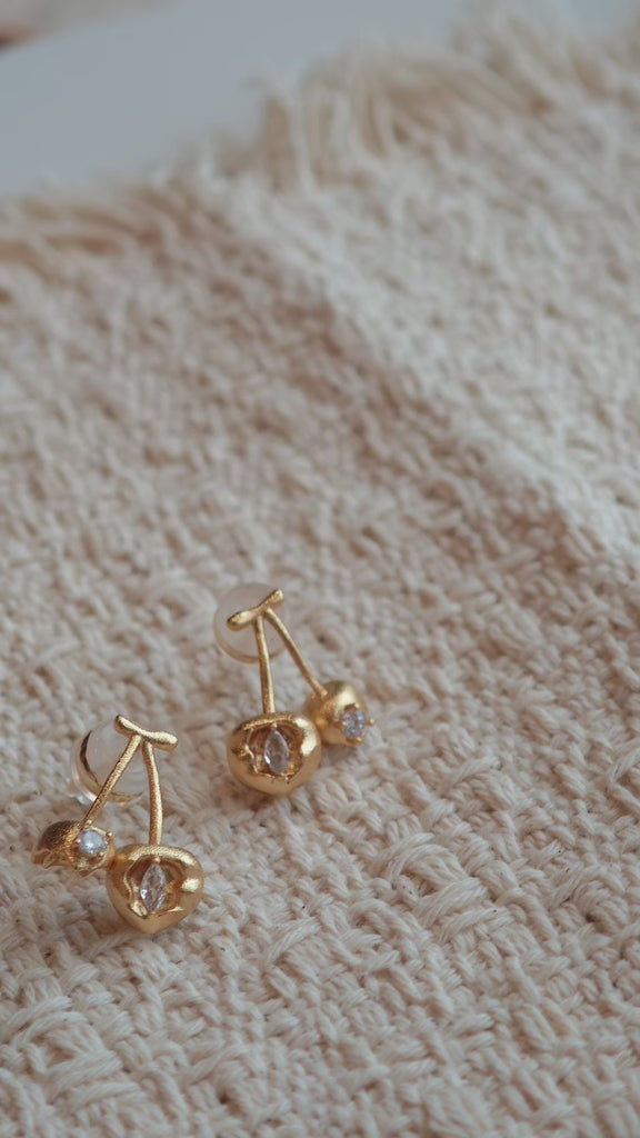 These cherry-shaped stud earrings feature a luxurious matte sandblasted finish, crafted from thick-plated real gold over 925 sterling silver posts. Each earring is designed as a pair of cherries, adorned with a marquise-cut and a round-cut sparkling clear stone, adding a delicate, eye-catching sparkle. The playful, organic cherry silhouette and premium gold plating make them a chic, elegant accessory for everyday wear.