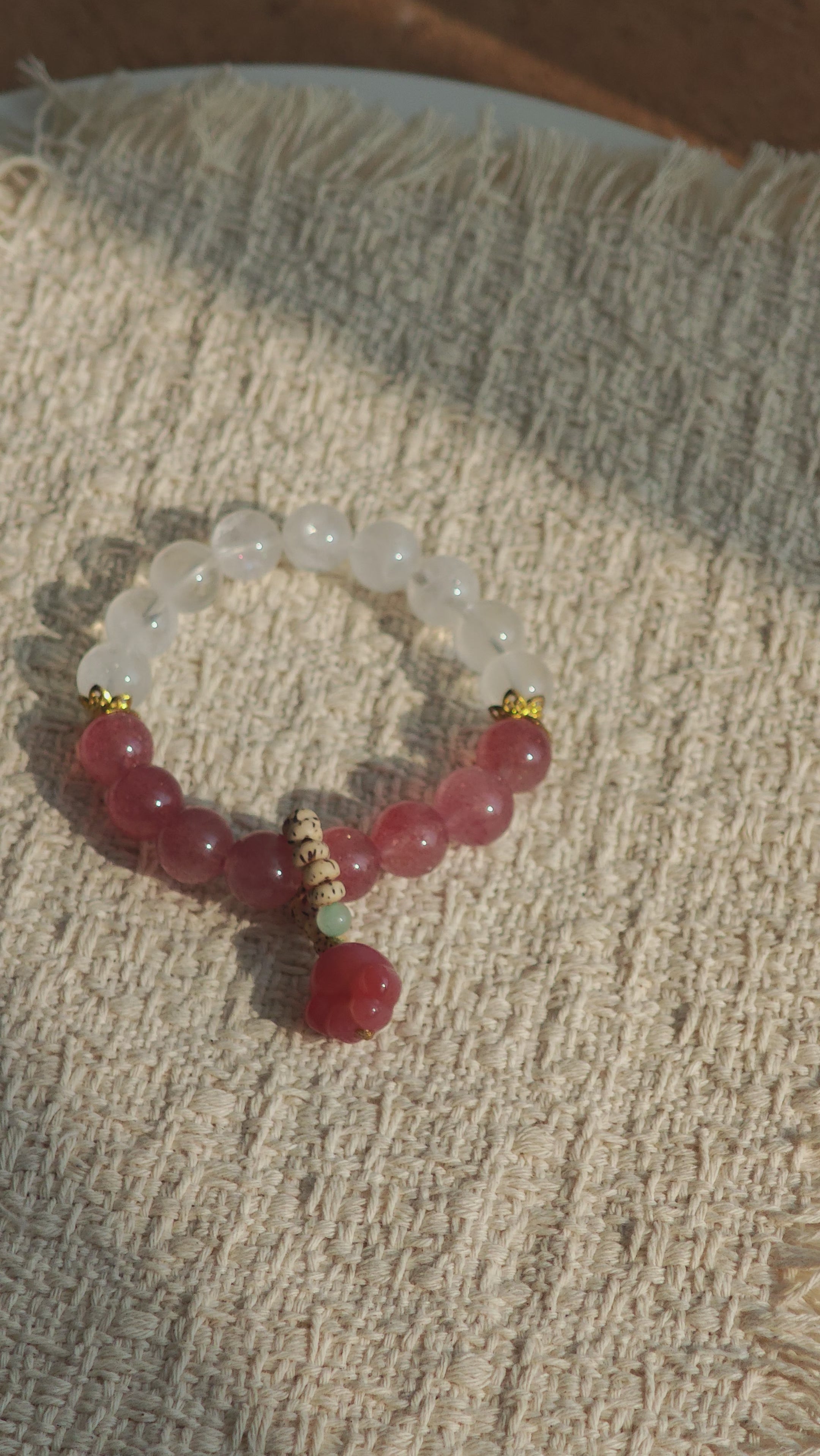 This bracelet features a beautiful blend of translucent white phantom quartz beads and rich pink strawberry quartz beads, creating a striking contrast. Adorned with delicate gold-toned accents and a charming red cat's paw charm, it exudes a sweet and elegant vibe. The combination of smooth crystal spheres and a playful pendant makes it a lovely and eye-catching accessory.