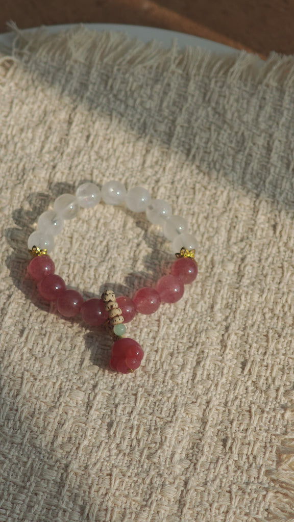 This bracelet features a beautiful blend of translucent white phantom quartz beads and rich pink strawberry quartz beads, creating a striking contrast. Adorned with delicate gold-toned accents and a charming red cat's paw charm, it exudes a sweet and elegant vibe. The combination of smooth crystal spheres and a playful pendant makes it a lovely and eye-catching accessory.